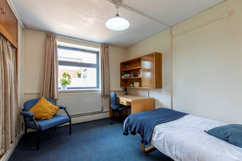 A room with single bed and a desk against one wall, and armchair and a wardrobe at the opposite wall. The desk as an office chair next to it and a set of shelves on the wall above it.