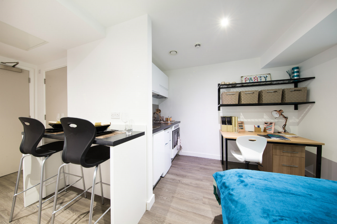 A photograph of a studio flat. Visible is the end of a bed, a kitchenette, a sideboard/table with chairs, and a desk with shelves above.