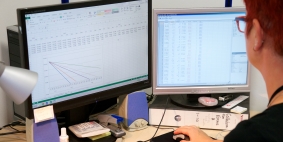 A person with red hair sat in front of two computer screens. Once has a graph on it and one has a spreadsheet on it