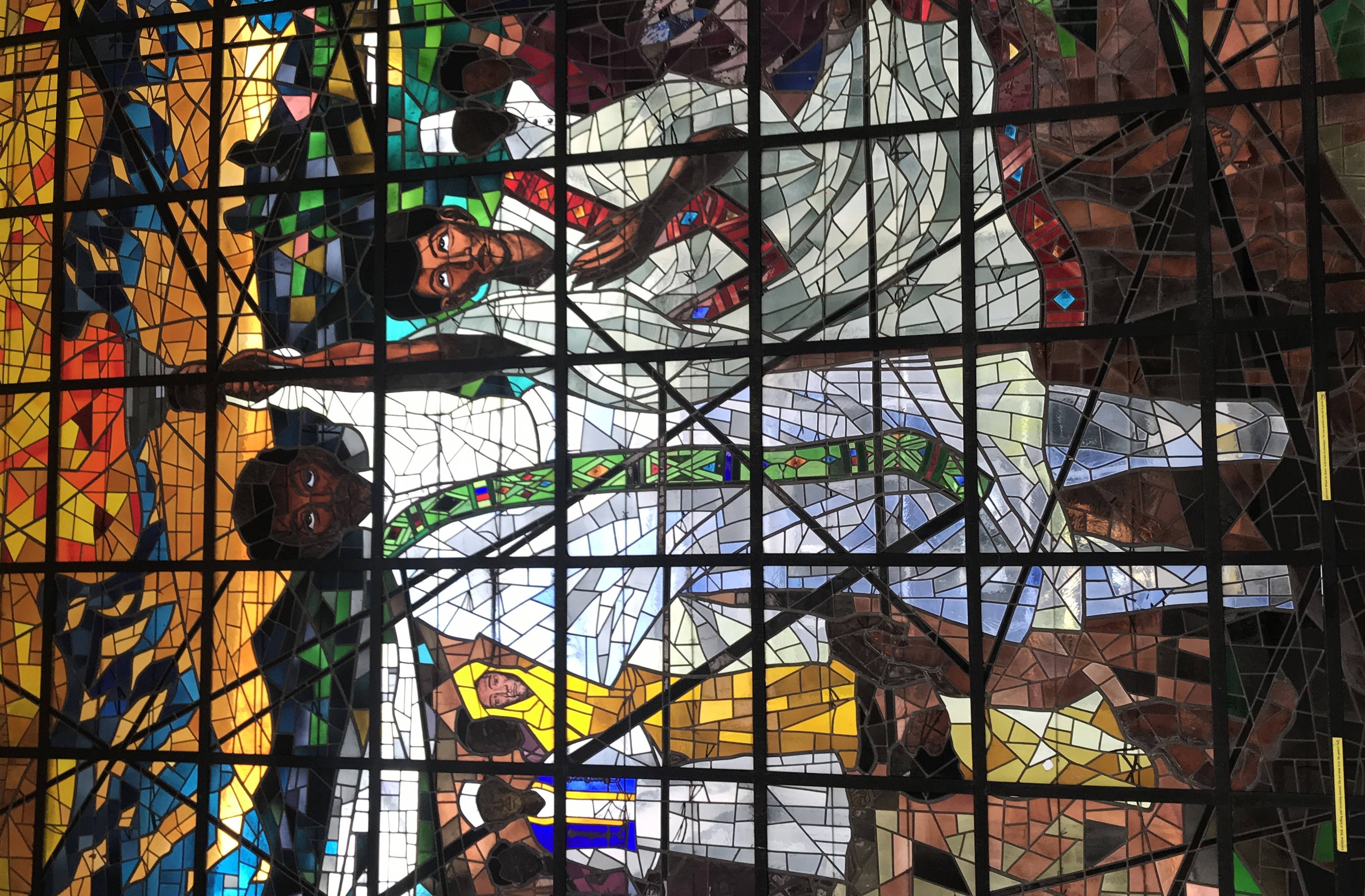 A stained glass window depicting a black man wearing white robes and colourful scarf (green background with diamond shapes of coloure in blues, purples and orange) holding a flaming torch. Beside him another black man wearing white robe with red hem showing diamond shapes of varying coloures. in the background are several people wearing colourful robes.