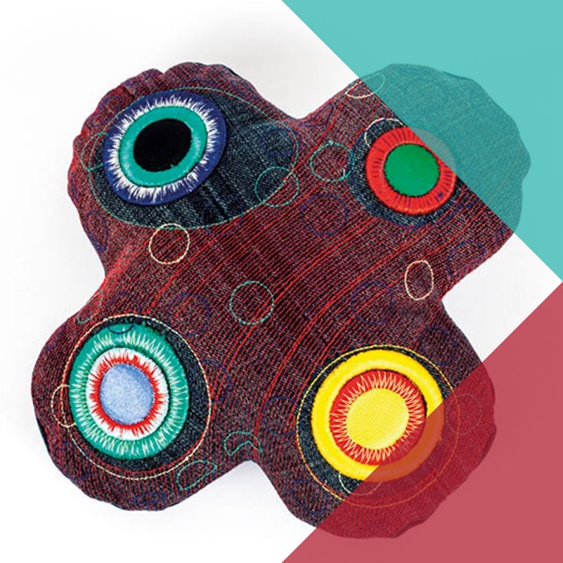 A photograph of a specially designed tactile cushion. The cushion is in the shape of an X with rounded edges and different shaped circles embroidered on each of its protrusions