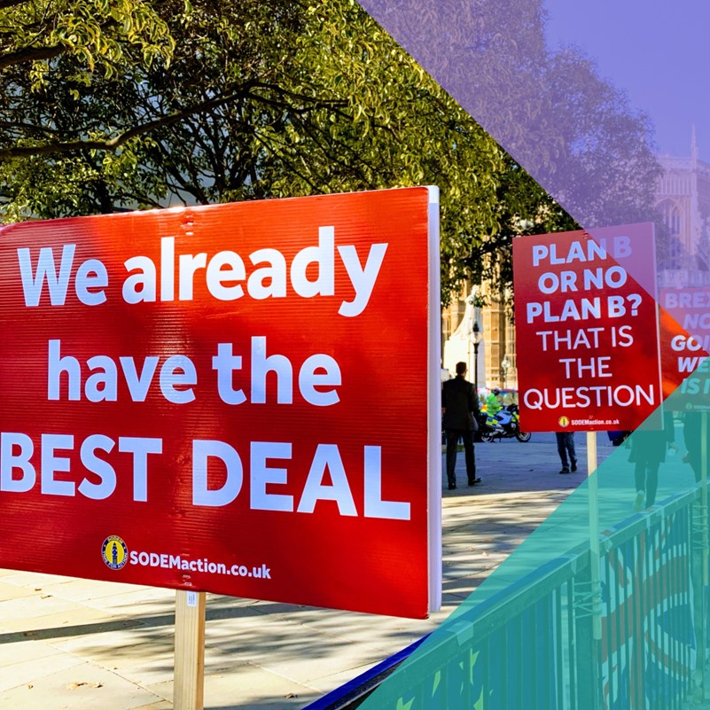 A photo of a street with a series of picket signs, one says 'We already have the BEST DEAL' another says 'PLAN B OR NO PLAN B? THAT IS THE QUESTION' and the third is only partially legible. There is a Union Jack next to an EU flag on the gate with the signs