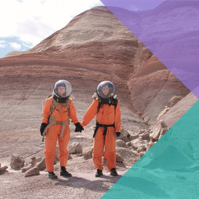 A photo of two women in orange space suits stood holding hands in an arid desert environment