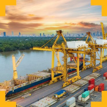 A photograph of shipping docks beside a large river with a forest behind and a city in the background