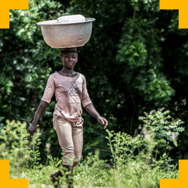 A young woman from Togo walking through a wooded area carrying a large container on her head