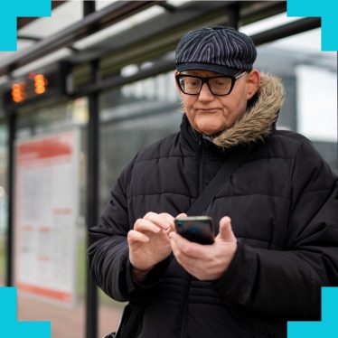 An older man stood at a bus stop holding out and using their smartphone