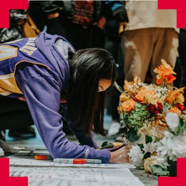 A woman kneeled down writing on a memorial card in front of flowers with people behind her in the background