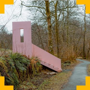 A photograph of a country road with a sculpture of a section of a house, a front door and set of stairs, on the side of the road.