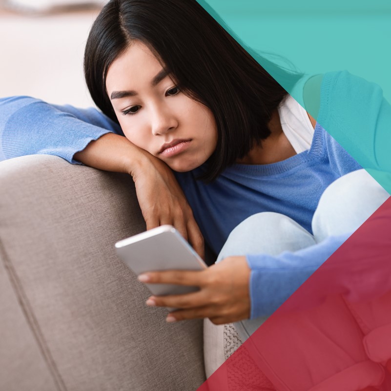 A photo of a teenage girl sat on a couch on her laptop with her phone beside her