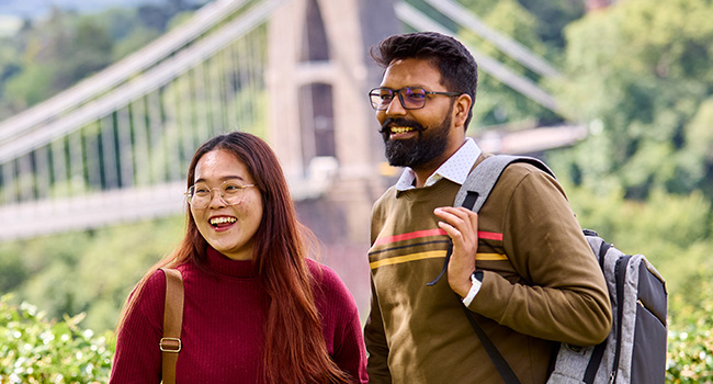 Image of a male student and a female student stood smiling in front of the Clifton Suspension Bridge