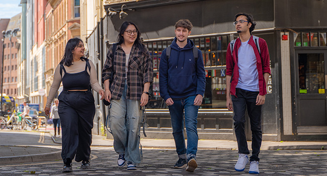 Four students walking and smiling in the centre of Bristol on a patterned brick floor with old buildings in the background