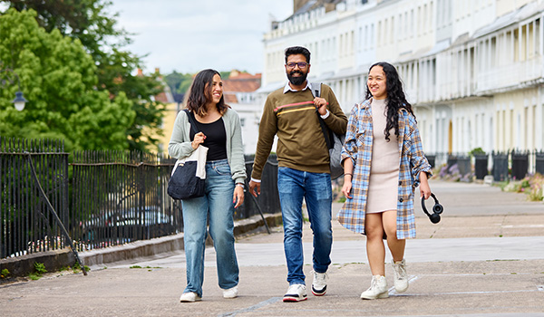 Three students walking along a scenic path in front of a curved row of historic houses in Bristol. The students are laughing and smiling as they walk, wearing casual clothing.