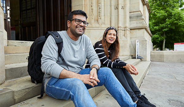 2 students sat on a step outside a historic campus building, looking into the distance and laughing