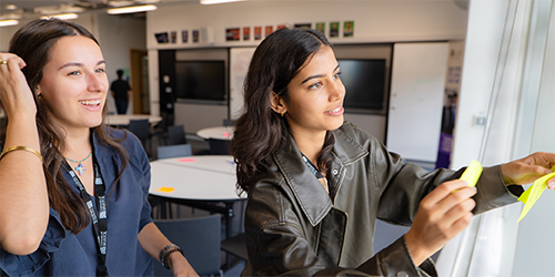 Two students putting sticky notes up on a board.