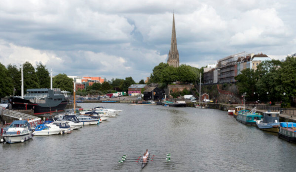 a wide river with many boats moored at the sides and a rowing crew in the middle. in the background is a parade of houses, trees and a large church spire.