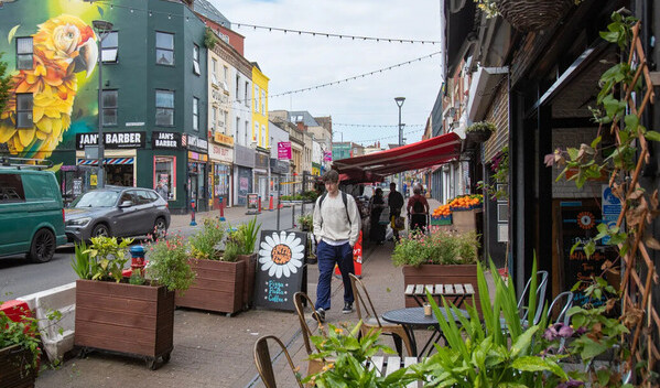 A young man walks down a high street with large pavement planters and greenery surrounding the cafe outdoor seating. a large yellow parrot is spray painted across a building in the background.
