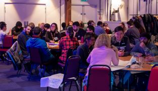 PhD students sitting at numerous round tables in a large room having discussions about PhD challenges 