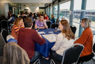 Six PhD students sitting around a round table in a large room engaged in discussion