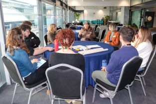 Ten people sitting around a round table listening to Pam Lock (centre) giving a facilitator briefingng