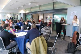 A large open plan room containing several round tables at which people are sat listening to a welcome talk