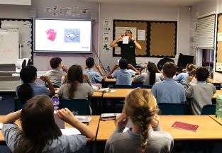 Dr Francesco Mitrotta blows across a sheet of paper to demonstrate lift while Year 6 pupils watch and carry out the same experiment at their desks.
