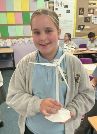 A Year 6 pupil holds up her completed paper wind turbine model in a classroom.