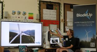 A presenter points to an image of a wind turbine on a large TV screen while pupils watch. Only the tops of the pupils' heads are visible.