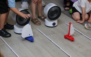 Two fans blow model sail cars along parallel tracks. The arms and legs of children setting off the cars and observing nearby are visible.