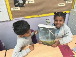 Two Year 6 pupils sit at a classroom desk, with one pupil smiling while holding up a Vestas paper wind turbine kit.