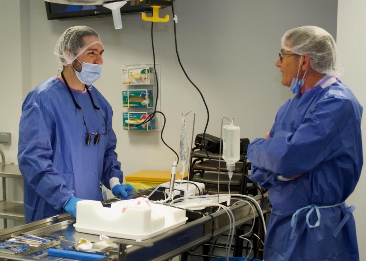 A student and lecturer in full scrubs stand over a lab table. They are discussing the sinus grafting practical procedure the student is currently undertaking. A motor is set up in background with a drip attached and instruments are in the foreground