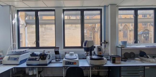 This is a photograph of a lab white lab bench, with large windows behind it. On the bench are two polishing machines, an auto grinder and a thin-sectioning machine.