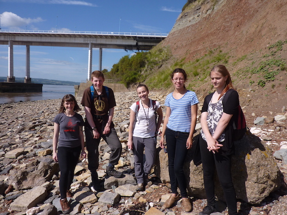 A group of five student interns standing on rocky terrain, enjoying a sunny day of field work on the Triassic-Jurassic boundary at Aust Cliff, near Bristol.