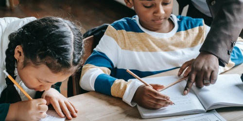 Two Black primary school children, one a girl and one a boy, sit writing calmly at a classroom desk. The Black teacher's hand reaches into the image to point out something on the boy's workbook.