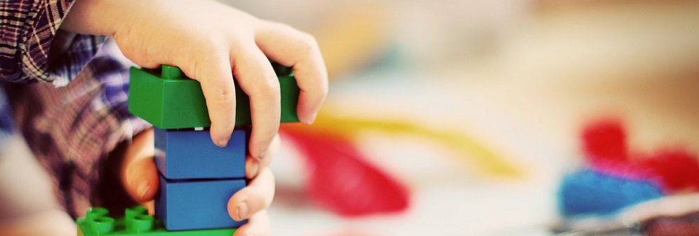 A child's hands as they build with brightly coloured blue and green plastic building blocks, with other toys in the background.