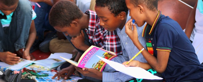 A group of Black primary school boys from Somalia sit in a group, with books, paper and pencils in front of them. The three boys in the middle of the image appear engrossed in working on a problem together.