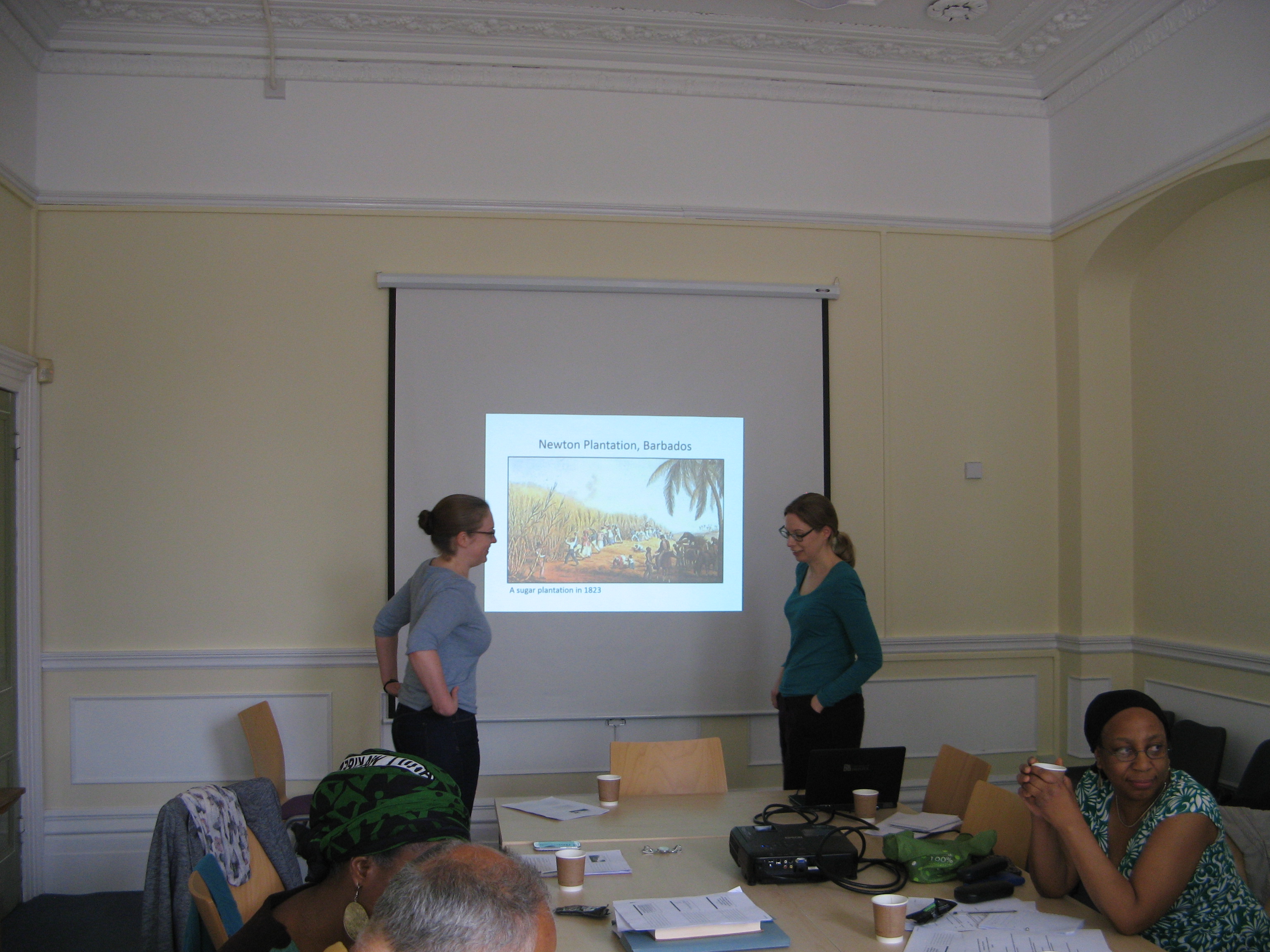 Photo of two women by a projector screen and participants sitting around table