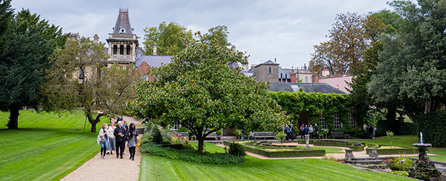 A group of people walking along a path which sits amongst a lush green lawn. To the right are trees and a water feature. Behind is and old, tall building.