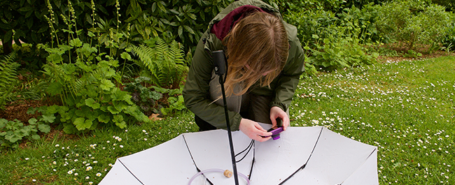 A person knelt down outdoors on grass. They are photographing objects on a white umbrella which is upside down