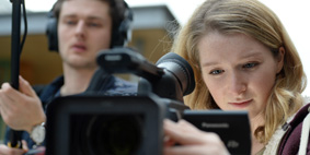 A female student looking through/focussing the lens of a camera, a male student stood beside her wearing headphones and holding a long-armed microphone.