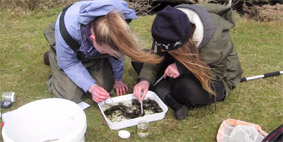 Two white women sat on grass and examining the contents of a white plastic container.