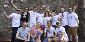A group of men and women standing and kneeling together and smiling. Many of them are wearing Pride t-shirts and holding rainbow flags.