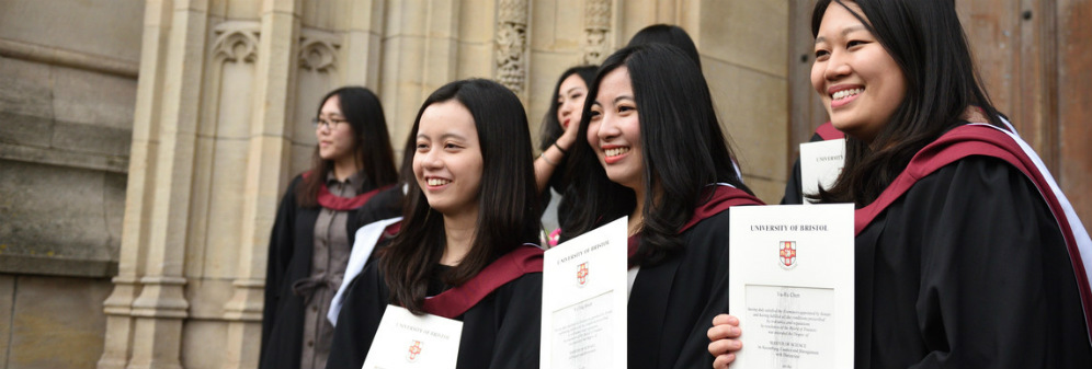 Three graduates standing outside Wills Memorial Building holding up their degree certificates.