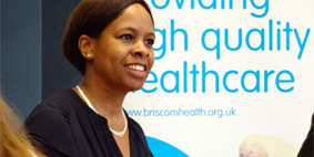 A black woman sitting and smiling in front of a sign that says 'Providing high quality healthcare, www.briscomhealth.org.uk'.