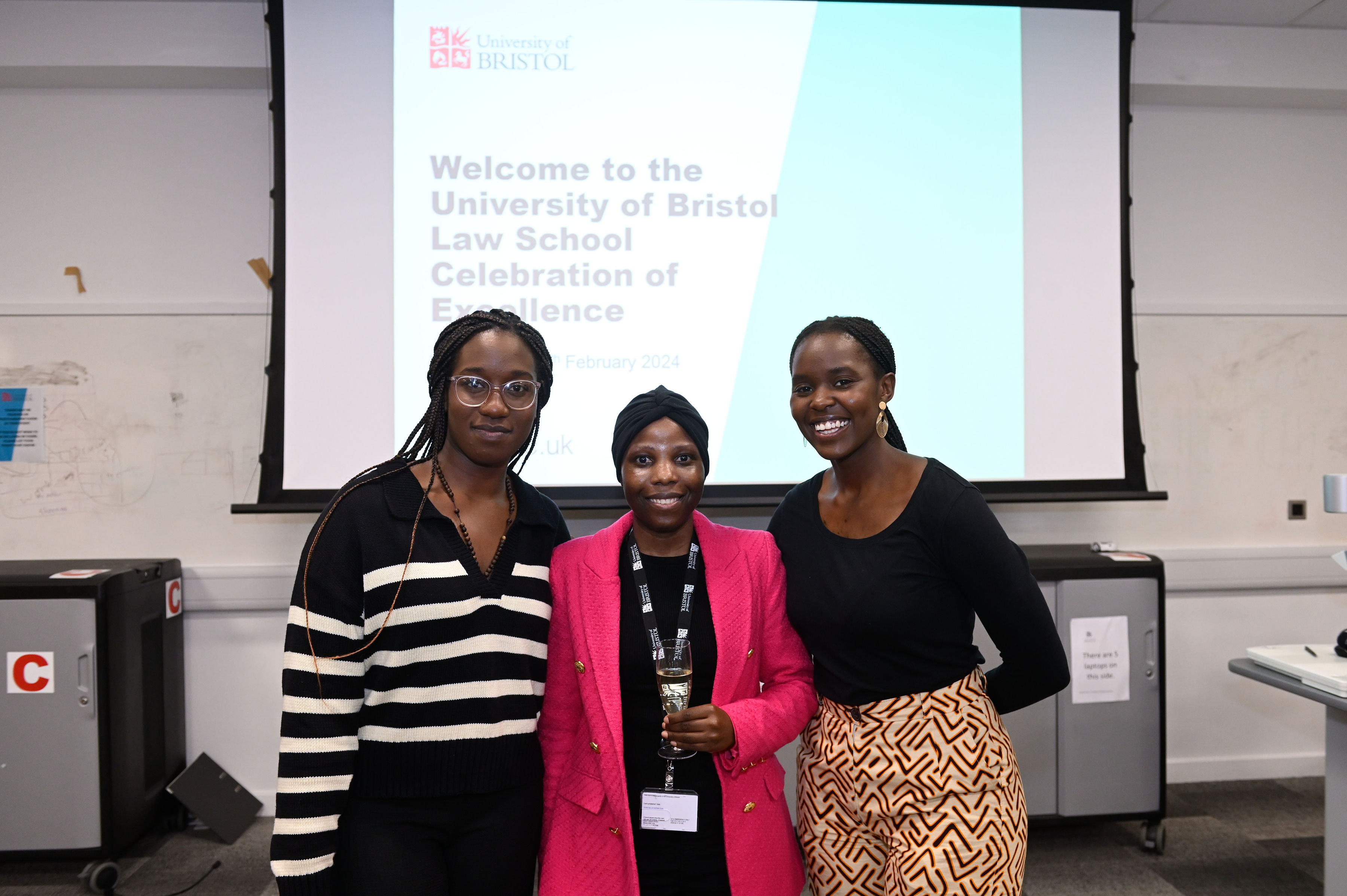 Group of thre law student female prize winners, stood in a row in front of a lecture room screen, smiling wearing smart clothes.