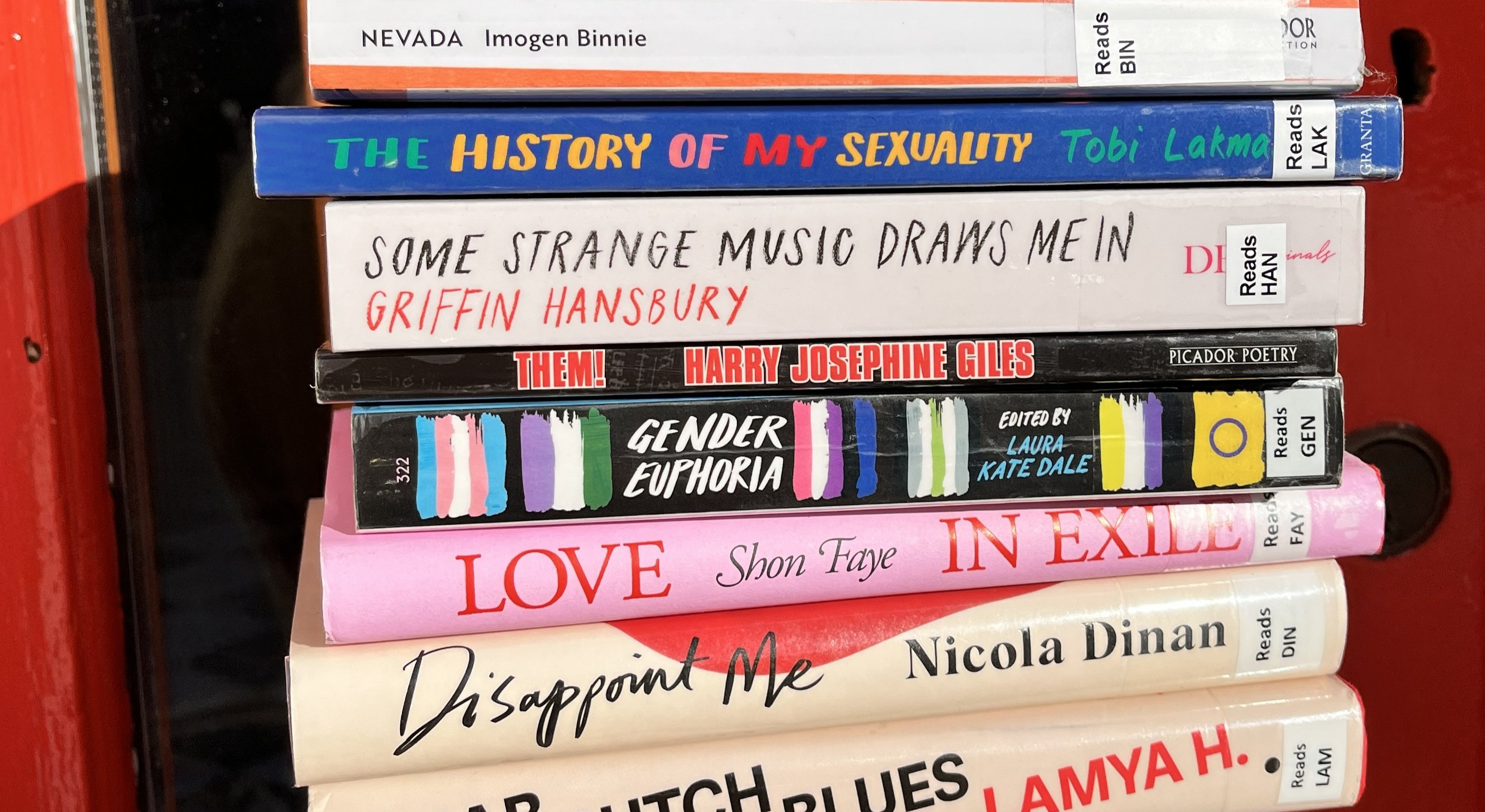 A stack of books about the LGBTQ+ community or by LGBTQ+ authors. Some titles include Love in Exile by Shon Faye, Disappoint Me by Nicola Dinan, and Nevada by Imogen Binnie. The book spines are standing colourful and proud.