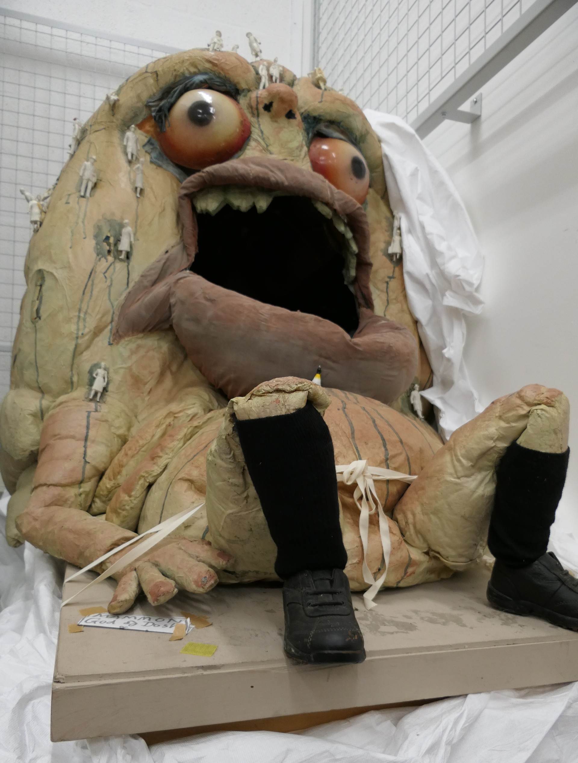 A large puppet sits on a table. He has big eyes and an open mouth and tiny figures of people are on his sides
