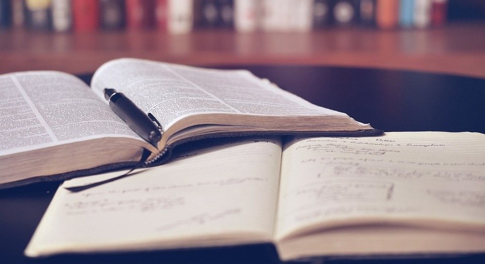 A desk with an open book, a notebook with writing in it and a pen. A bookshelf in the background.