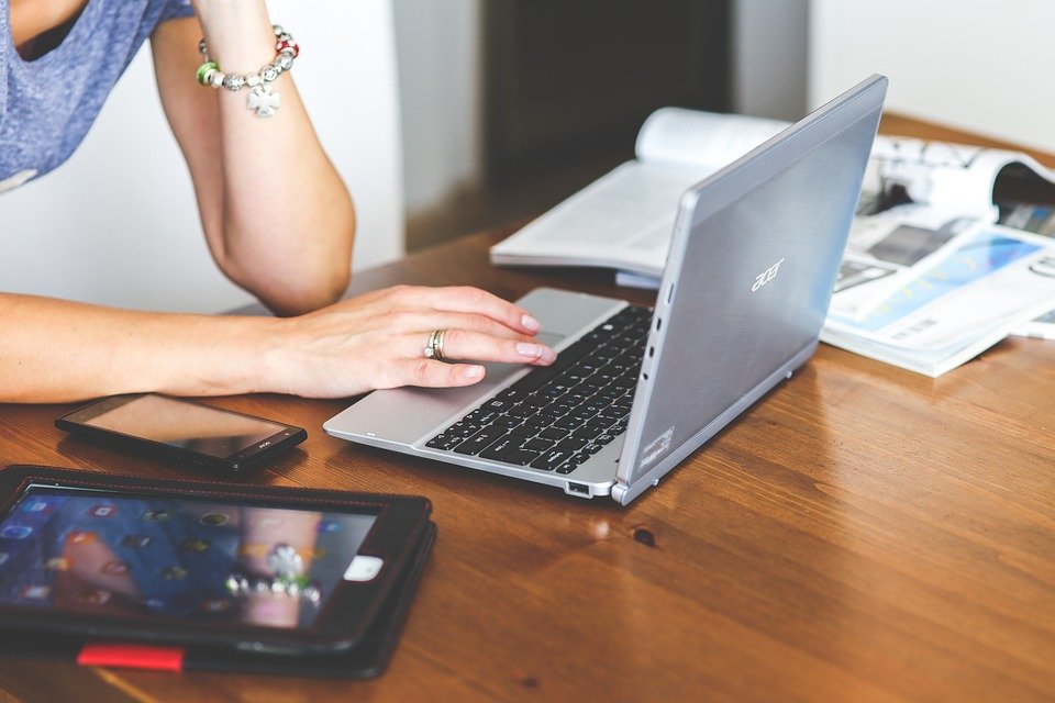 Desk with laptop, phone and tablet as well as open books. Students arms with jewellery are visible working on the laptop. 