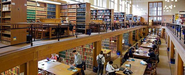 Shows one of the wings of the Wills Memorial Library. A large wood panelled library on two floors, with book shelf alcoves with large study desks and lots of students studying.