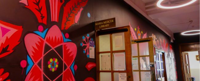 Entrance to the physics library. A corridor is painted vibrant multicolours with physics inspired illustrations like atoms. This leads to the old wooden doors of the library. A vintage sign above the doors reads 